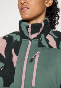 Donna che indossa una giacca in pile verde e rosa con motivo camouflage, colletto alto e due tasche con zip rosa sul petto.