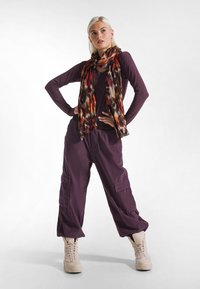 Camiseta de manga larga morada, pantalones cargo con grandes bolsillos y una bufanda de colores rojo y marrón con estampado. Botas beige con cordones. Fondo blanco liso.