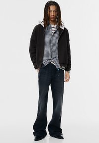 Giacca nera con cappuccio sopra una camicia a quadri, jeans scuri con gambe svasate e scarpe nere eleganti. Presenta texture e colori stratificati.