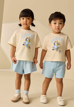 Zwei Kleinkinder tragen passende beige T-Shirts mit einem Cartoon-Hund und gestreifte blaue Shorts und stehen nebeneinander.