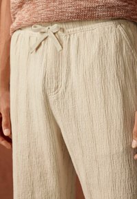 Beige teksturerede bukser med elastisk talje og snørelukning, med sidelommer og lodrette linjer i stoffet.