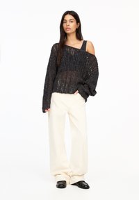 Czarny sweter dzianinowy o luźnym, otwartym splotzie, szerokich rękawach i dekolcie off-shoulder, w połączeniu z beżowymi spodniami o szerokich nogawkach i czarnymi butami typu slip-on.