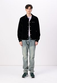 Black corduroy jacket with snap buttons, shirt collar, and white logo detail, paired with light blue jeans and green athletic shoes.