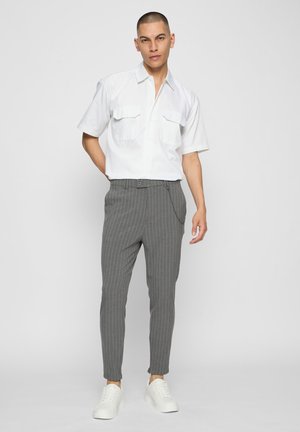 Vyras dėvi baltą trumpomis rankovėmis mygtukais užsegamą marškinius, pilkus dryžuotus kelnes su grandinėlės detale ir baltus sportinius batelius, stovintis prieš paprastą šviesų foną.