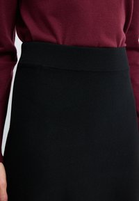 Zwarte kokerrok met een geribbelde tailleband en een gladde textuur; gecombineerd met een bordeauxkleurig langwerpig shirt in een close-up weergave.