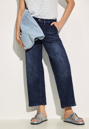 Osoba ubrana w ciemnoniebieskie szerokie jeansy, bezrękawnik w paski, trzyma jasnoniebieską kurtkę, stoi w srebrnych sandałach z klamrą.