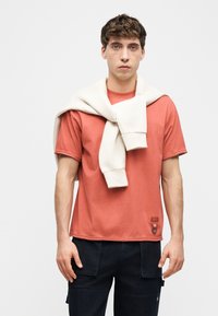 Νέος άνδρας φορώντας κοραλί μπλουζάκι και σκούρο παντελόνι με λευκή φανέλα περασμένη στους ώμους του, στέκεται μπροστά σε απλό φόντο.