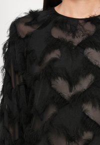 Haut en tissu noir transparent avec des motifs texturés et duveteux en forme de losanges couvrant la poitrine et les épaules.