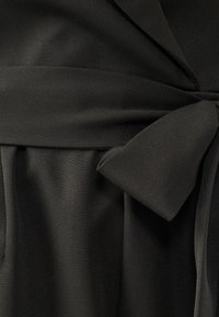 Black fabric garment with a smooth texture, featuring a tied sash at the waist and pleated details along the bottom.