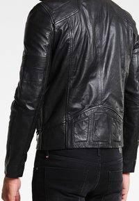 Schwarze Leder-Motorradjacke mit strukturierten Einsätzen, sichtbaren Nähten und tailliertem Design. Reißverschluss vorne, minimale Hardware.