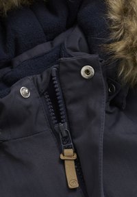 Mörk marinblå jacka med en strukturerad tyg, mjukt fleecfoder, dragkedja och tryckknappar, samt en huva med pälsfoder.