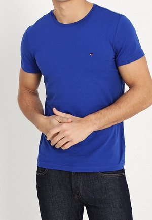 Mand iført en tætsiddende kongeblå T-shirt med et lille logo på brystet og mørke jeans står mod en ensfarvet hvid baggrund.