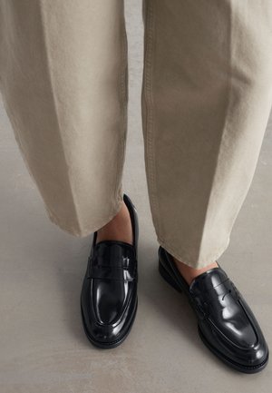Personne portant un pantalon beige et des mocassins en cuir noir brillant, debout sur un sol lisse gris clair.