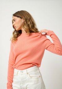 Licht koraalroze sweater van zacht materiaal, met lange mouwen en een ronde halslijn. Gecombineerd met witte high-waisted jeans.