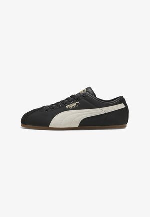 Baskets Puma noires à profil bas avec une bande latérale beige, des lacets noirs et une semelle en gomme, montrant le côté extérieur gauche avec le logo "Puma Tackle".