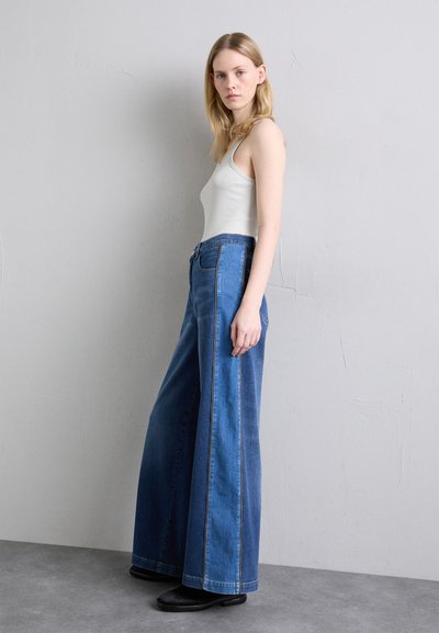 Weite blaue Jeans aus Denim mit auffälligen gelben Nähten, hoher Taille und Fünf-Taschen-Design, kombiniert mit einem hellgrauen Tanktop.