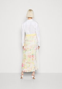 Frau mit kurzem blonden Haar, die ein weißes Shirt und einen plissierten, floral gemusterten Rock mit gelbem Bund trägt, vor einem schlichten weißen Hintergrund steht.