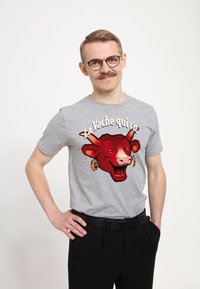 Šedé bavlněné tričko s červenou grafikou kreslené krávy s náušnicemi a textem "La Vache qui rit." Kombinováno s černými kalhotami.