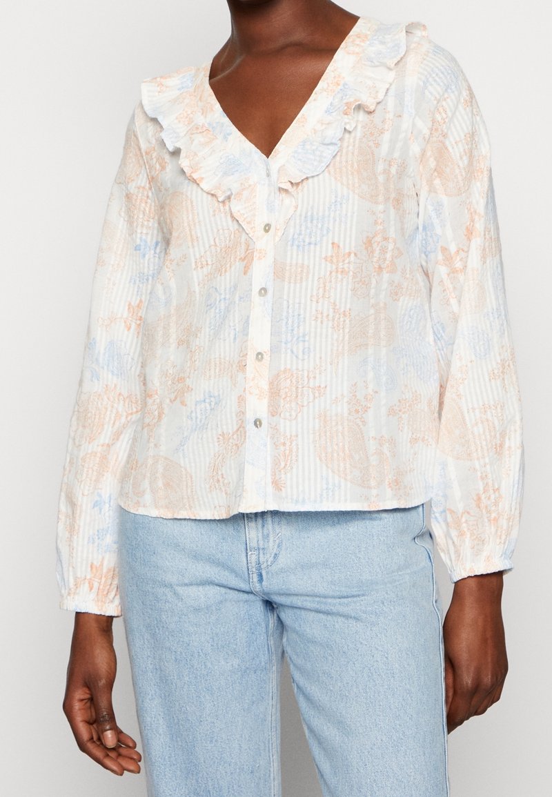 Femme portant un chemisier blanc boutonné avec des motifs floraux pastel, col en V à volants et manches longues, associé à un jean bleu clair.