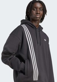 Svart hettegenser med hvitt tre-stripes design på venstre side. Har en frontlomme, snor til hetten, og lite logo på brystet.