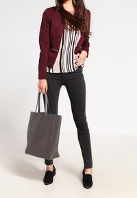 Veste bordeaux zippée portée sur un haut rayé, associée à un jean noir et des chaussures noires. Sac fourre-tout gris avec un détail de couture minimaliste.