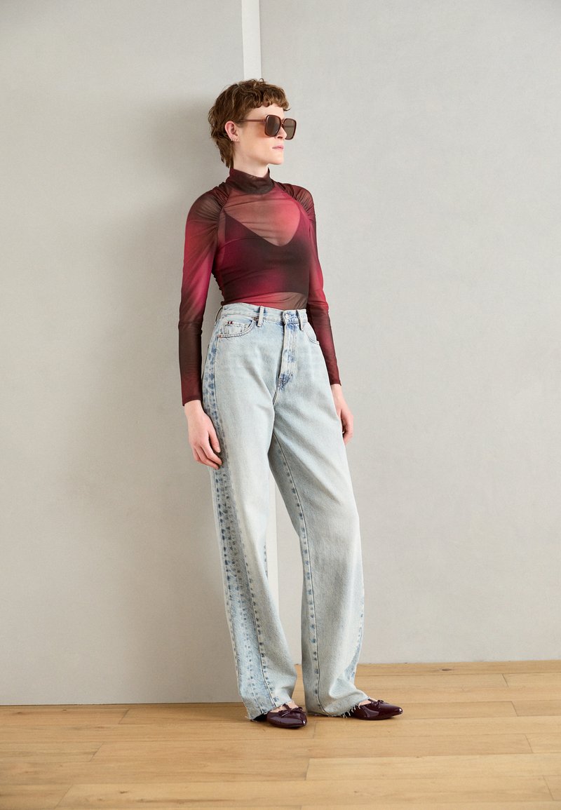 Top trasparente a maniche lunghe in gradiente dal rosso al marrone, abbinato a jeans baggy a vita alta di colore azzurro chiaro. Scarpe a punta color viola scuro.