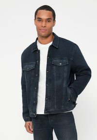 Muškarac u tamnoplavoj traper jakni preko bijele majice, s jednom rukom u džepu, stoji naslonjen na bijelu pozadinu.