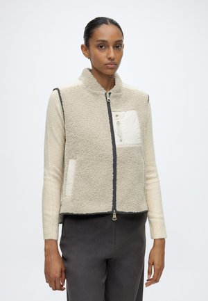 Femme portant une veste polaire crème zippée avec une poche poitrine, par-dessus un pull beige à manches longues et un pantalon foncé sur fond blanc.