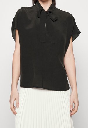 Femme portant un chemisier noir sans manches avec un nœud noué au cou et une jupe plissée blanche, debout devant un fond clair uni.