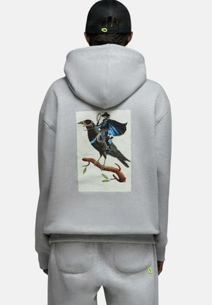 Personne portant un sweat à capuche gris avec un graphisme représentant un cow-boy chevauchant un grand oiseau noir perché sur une branche d'arbre dans le dos.