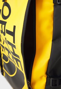 Zaino giallo e nero con un esterno durevole, dotato di una cerniera superiore e un grande logo frontale. Tessuto texturizzato e spallacci regolabili.