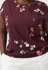 Bordeaux bloemenbloes met korte mouwen, een ronde halslijn en delicate witte en roze bloemenpatronen tegen de donkere achtergrond.