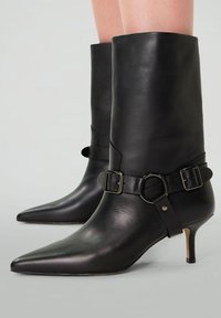 Bottines en cuir noir à bout pointu, petit talon et une sangle décorative avec boucle autour de la cheville. Texture lisse avec un design épuré.