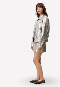 Ensemble veste et jupe en argent métallique, avec une texture lisse, une coupe décontractée et de grandes poches avant. Chaussures plates noires avec des accents argentés.