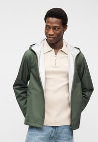 Νέος άνδρας που φοράει μπεζ μπλούζα πόλο με φερμουάρ, ανοιχτό πράσινο μπουφάν με κουκούλα και ανοιχτό μπλε τζιν, κοιτάζει ελαφρώς προς το πλάι με τα χέρια πίσω από την πλάτη.