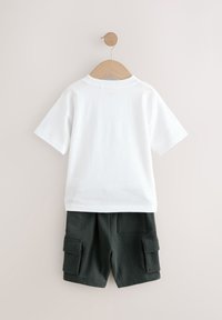 T-shirt en coton blanc et short cargo vert foncé suspendus à un crochet en bois, avec deux poches latérales sur le short.