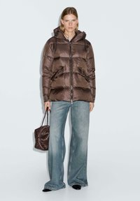 Donna che indossa una giacca imbottita marrone, jeans chiari a gamba larga, scarpe nere e tiene in mano una borsa texture marrone, in piedi contro uno sfondo semplice.
