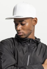 Cappello bianco con visiera piatta e cuciture sottili, indossato con una giacca nera con zip che presenta un cappuccio e polsini elastici.