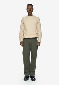 Sort mand iført en beige sweater, olivenfarvede bukser med et sort bælte og sorte kraftige støvler, stående fremadskuende mod en hvid baggrund.