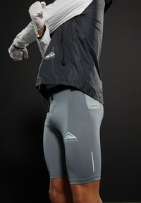 Shorts de sport gris avec un design ajusté. Présente une petite poche, un logo vert clair et des accents réfléchissants. Tissu texturé visible.