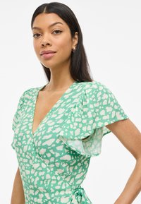 Grønt omslagkjole med hvitt abstrakt mønster, med bølgende ermer og bånddetalj i midjen. Lett, teksturert stoff.