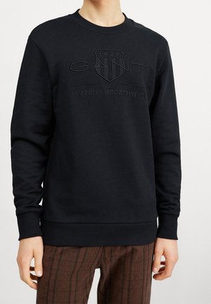 Person trägt einen schwarzen Pullover mit dem geprägten "GANT American Sportswear 1949"-Logo, kombiniert mit braunen gestreiften Hosen.
