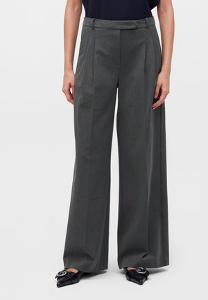 Personne portant un pantalon gris taille haute, à jambes larges, avec des plis sur le devant et des chaussures noires pointues avec détail de boucle argentée.