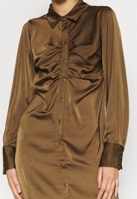 Brązowa satynowa sukienka koszulowa z kołnierzykiem, marszczeniem z przodu i zapięciem na guziki. Długie rękawy z guzikami przy mankietach.