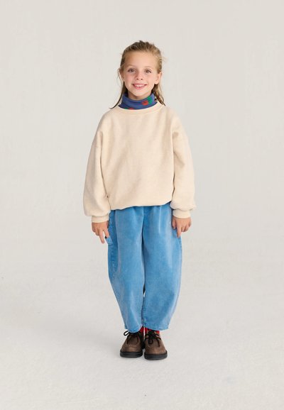 Jeune fille debout et souriante portant un pull beige, un pantalon bleu, un col roulé coloré et des chaussures marron sur un fond clair uni.