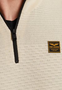 Beige, gestructureerde zip-up sweater met een zwarte rits en een PME Legend merkpatch met gele letters op de linkerkant van de borst.