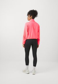 Veste cropped rose vif avec un col haut, associée à des leggings noirs et des baskets légères. Présente des poignets élastiques et une texture lisse.