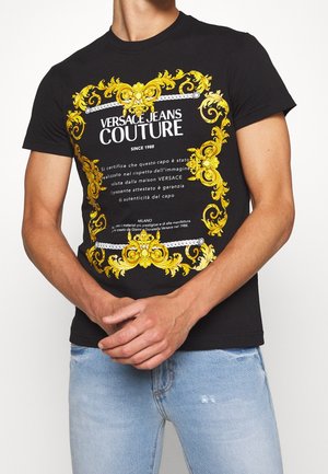 Czarny t-shirt z ozdobnym złotym wzorem graficznym i tekstem. Tkanina jest gładka, z okrągłym dekoltem. Noszony z jasnoniebieskimi, przetartymi dżinsami.