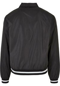 Chaqueta bomber negra con cuello tipo camisa, puños y dobladillo elásticos, que presenta rayas blancas en el dobladillo y los puños. Textura de tela suave.