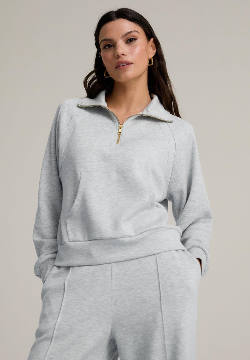 Ljusgrå zip-up sweatshirt med hög krage, raglanärmar, framficka och en mjuk textur. Guldiga dragkedjor som detaljer.